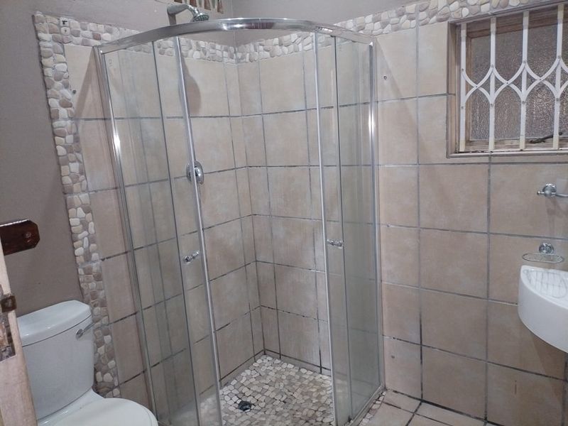 A functional bathroom featuring a corner shower enclosure, wall-mounted basin, and neutral ceramic tiling with decorative pebble accents.