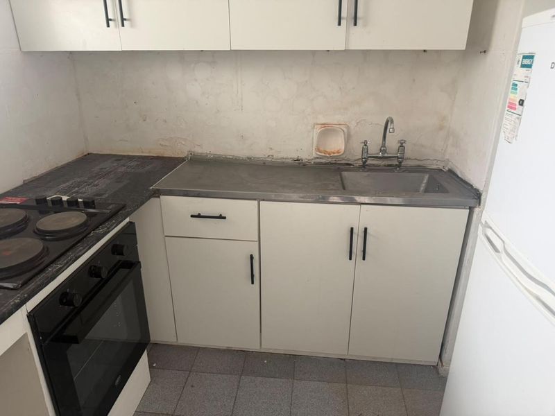 A compact kitchen featuring built-in cabinetry, an electric hob, oven, and stainless steel sink, currently requiring cosmetic updates and wall repairs.