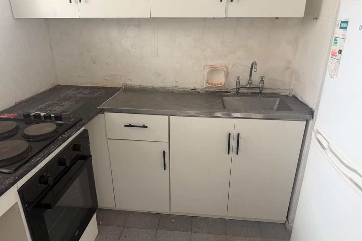 A compact kitchen featuring built-in cabinetry, an electric hob, oven, and stainless steel sink, currently requiring cosmetic updates and wall repairs.