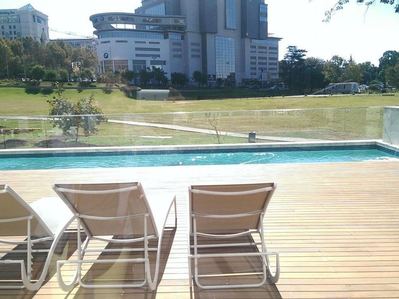 Outdoor pool area featuring timber decking, a glass balustrade, and a water feature, overlooking a landscaped park and urban skyline.