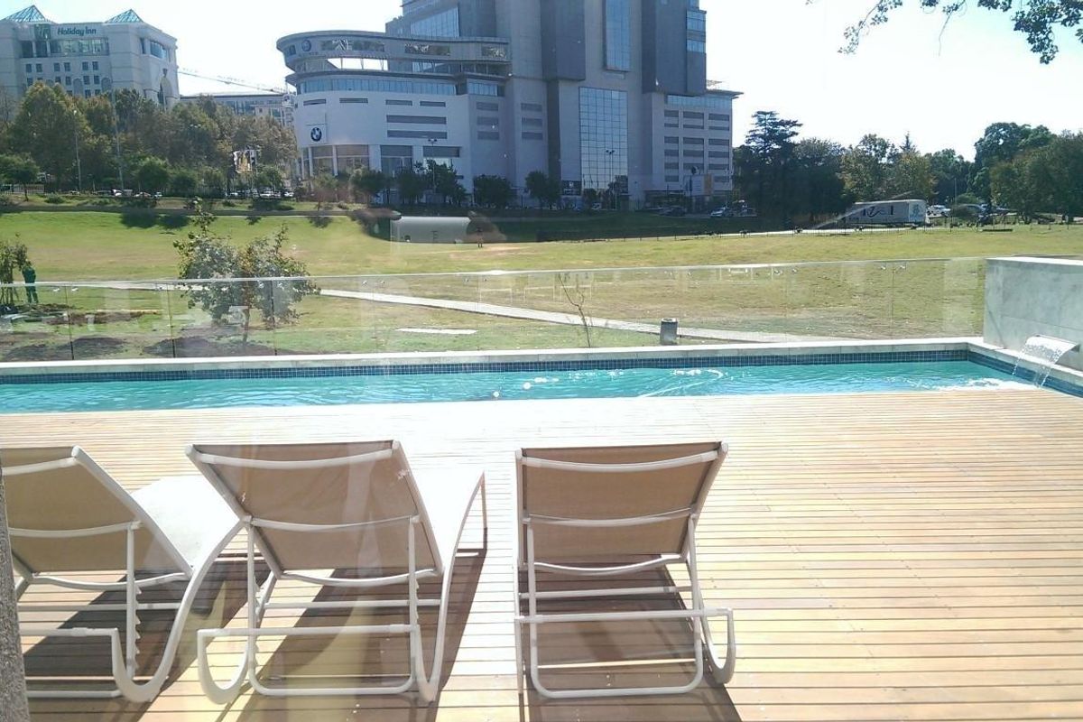 Outdoor pool area featuring timber decking, a glass balustrade, and a water feature, overlooking a landscaped park and urban skyline.