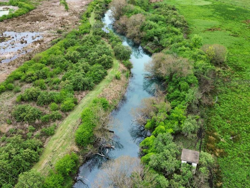Aerial view of a natural river landscape featuring lush vegetation, open green fields, and a small utility structure along the water bank.