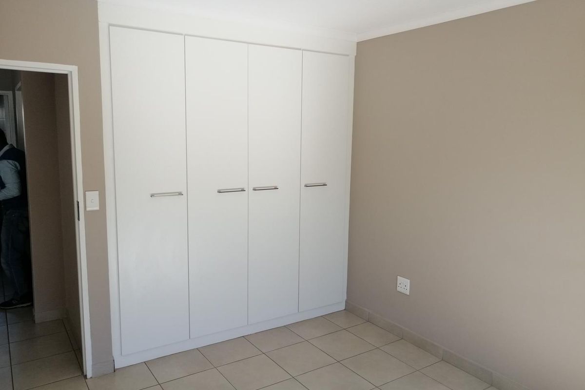 A clean, empty bedroom featuring neutral wall tones, tiled flooring, and modern white built-in cupboards with sleek handles.