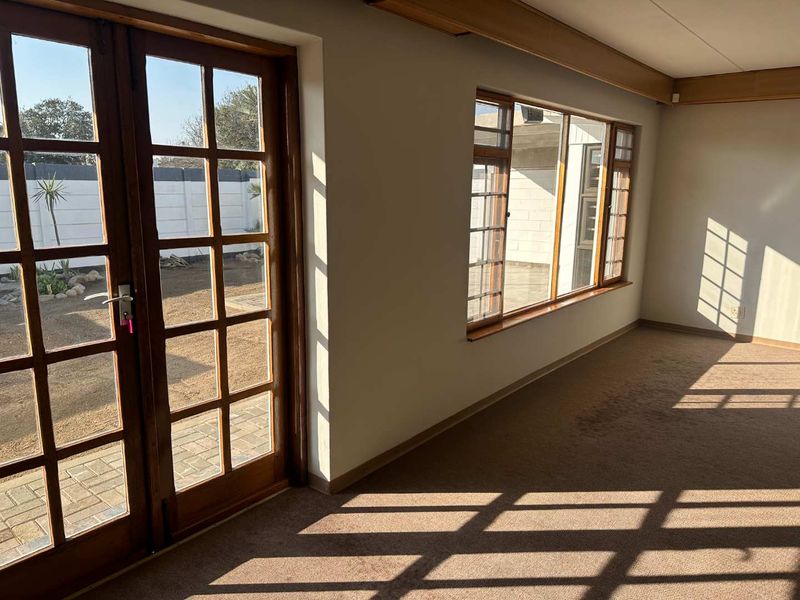 A bright, empty living room featuring timber-framed french doors and windows, carpeted flooring, and exposed timber ceiling beams, offering direct access to the garden.