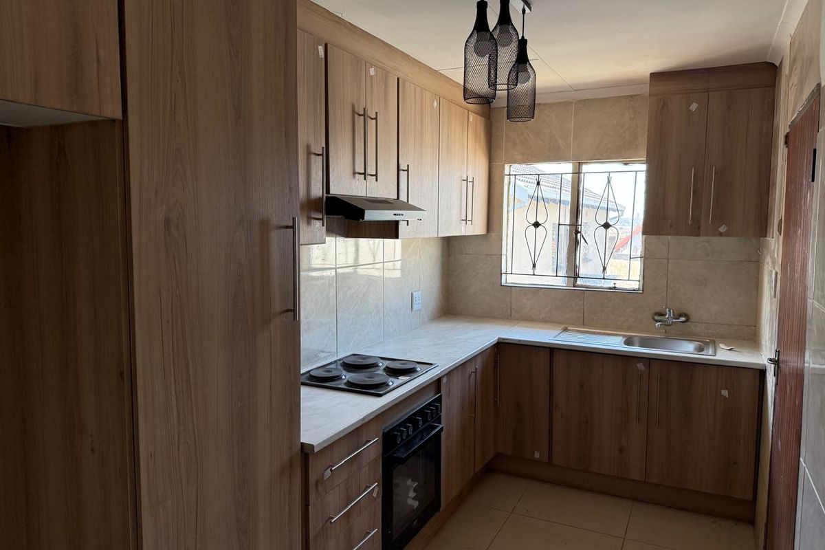 A functional kitchen featuring wood-look cabinetry, integrated electric hob and oven, tiled backsplash, and a stainless steel sink with security-barred window.
