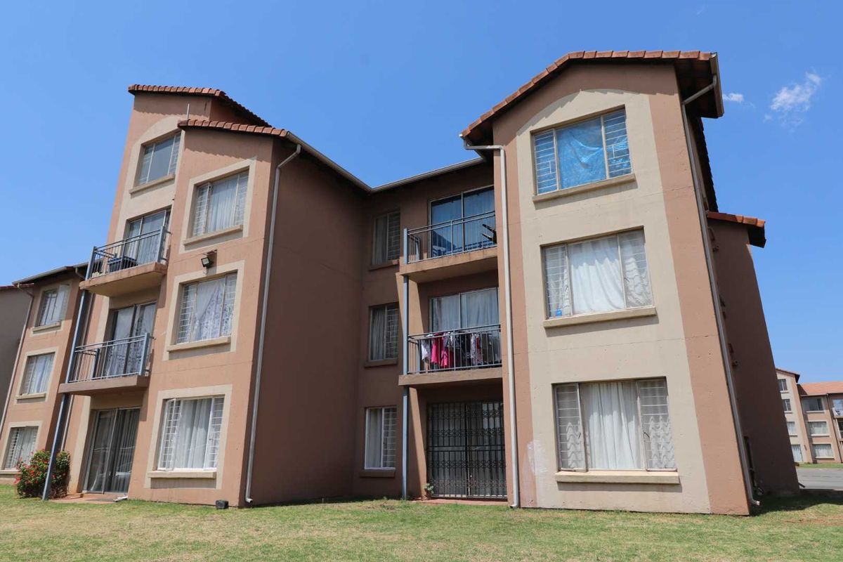 Exterior view of a multi-story apartment building featuring balconies, security gates, and tiled roofing set against a clear blue sky.
