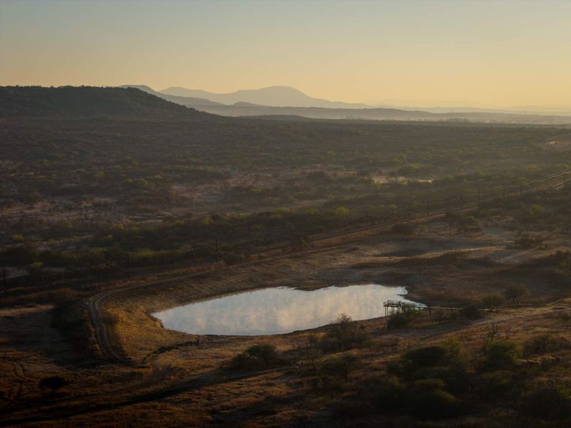 A scenic aerial view of a natural pond with a wooden viewing deck, set against a vast landscape of rolling hills and mountains.
