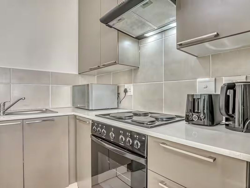 A modern, compact kitchen featuring sleek grey cabinetry, a built-in oven with electric hob, extractor fan, and a clean tiled backsplash.