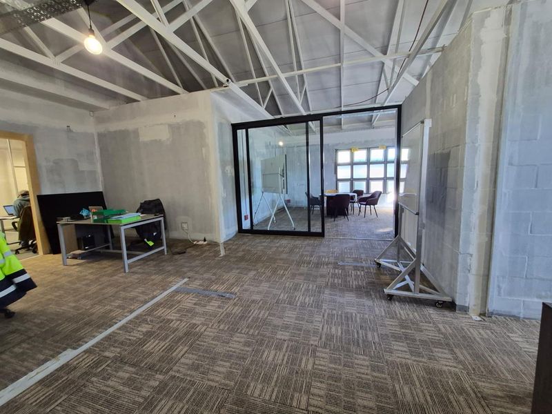 Open-plan office space featuring exposed steel roof trusses, carpet tiles, and a modern glass partition wall, currently in an unfinished shell condition.