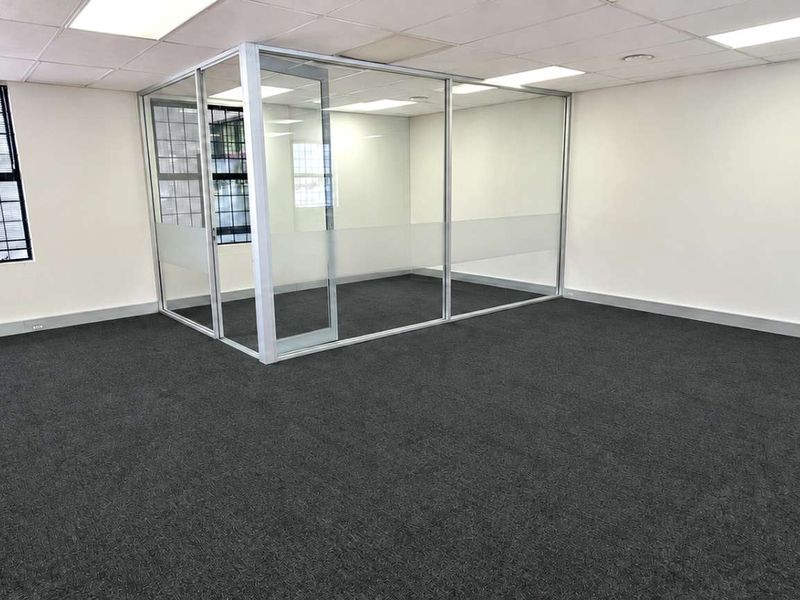 A bright, open-plan office space featuring modern glass partitioning, neutral walls, dark carpeting, and a suspended ceiling with integrated lighting.