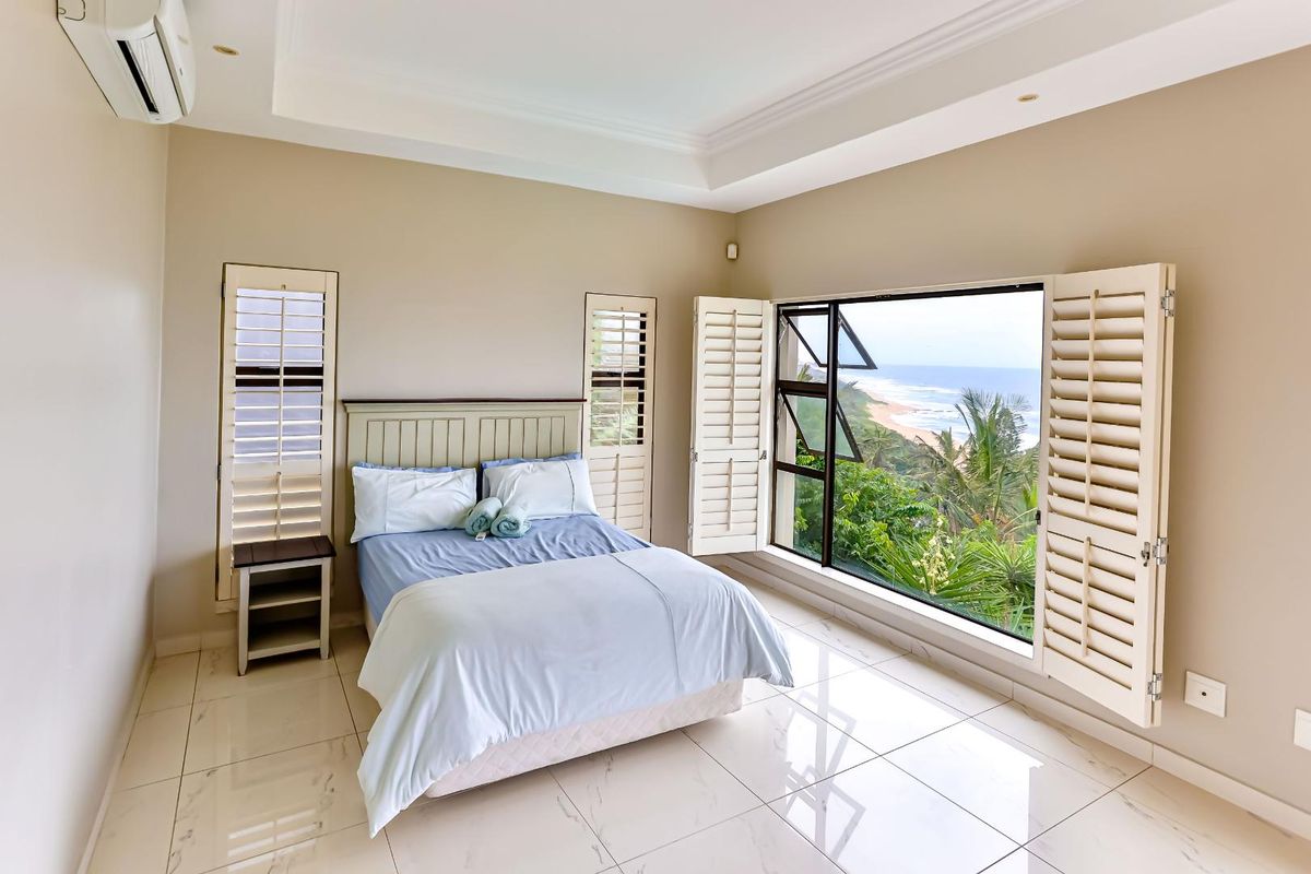 A bright bedroom featuring tiled flooring, plantation shutters, and a tray ceiling, offering expansive ocean views through large windows.