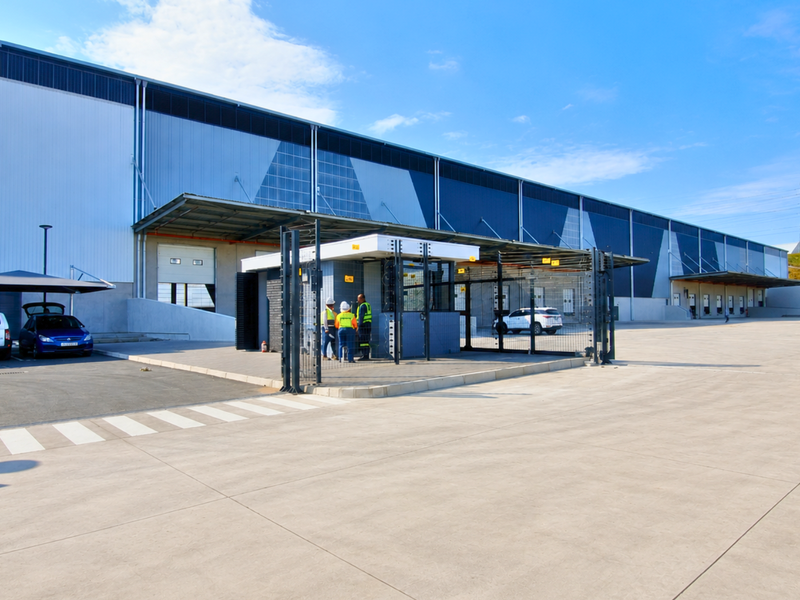 Modern industrial warehouse facility featuring a large concrete yard, secure guard house, multiple loading docks with roller shutter doors, and covered parking bays.