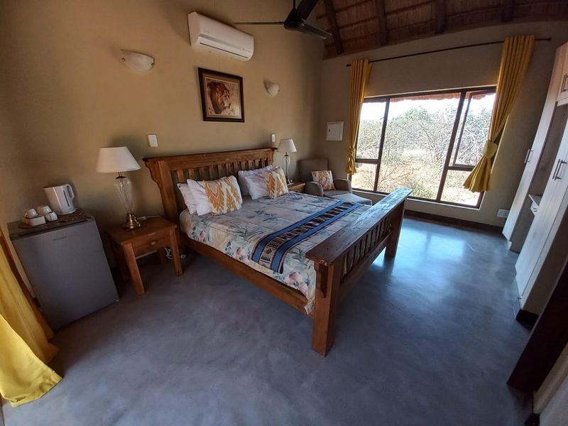 A spacious bedroom featuring a high thatched ceiling, polished concrete floors, air conditioning, and large windows with natural bushveld views.