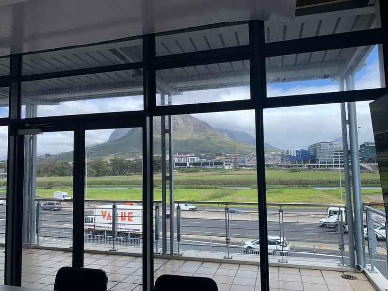 Modern office space featuring expansive floor-to-ceiling windows with panoramic mountain and highway views, leading out to a tiled balcony with a glass balustrade.