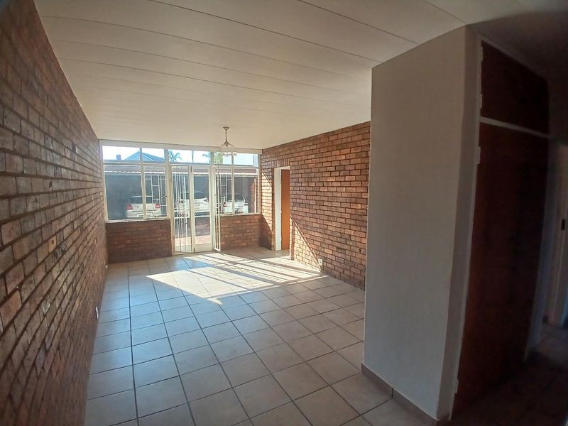 An empty living area featuring exposed brick walls, ceramic tiled flooring, and a built-in cupboard, with natural light entering through large windows.