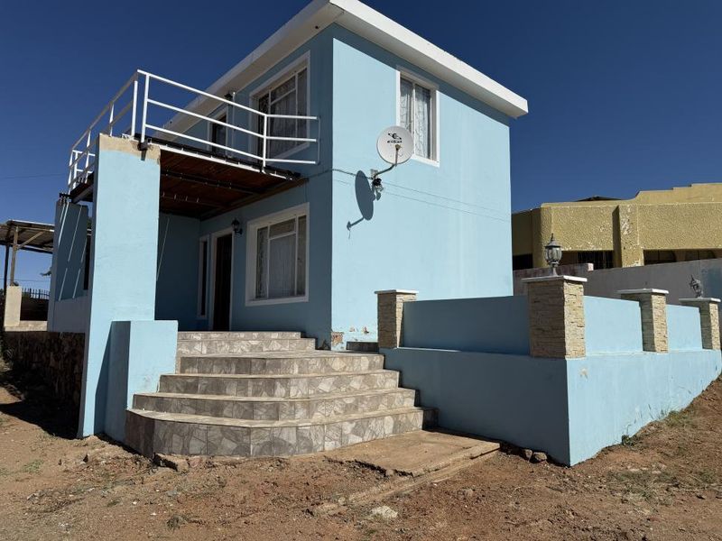 Front elevation of a two-story house featuring a tiled entrance staircase, a balcony with metal railings, and a low boundary wall with stone-clad pillars.
