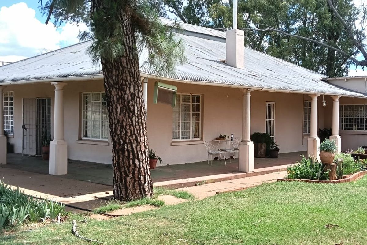 A traditional farmhouse featuring a wrap-around veranda with classic columns, corrugated iron roofing, and established garden surroundings.