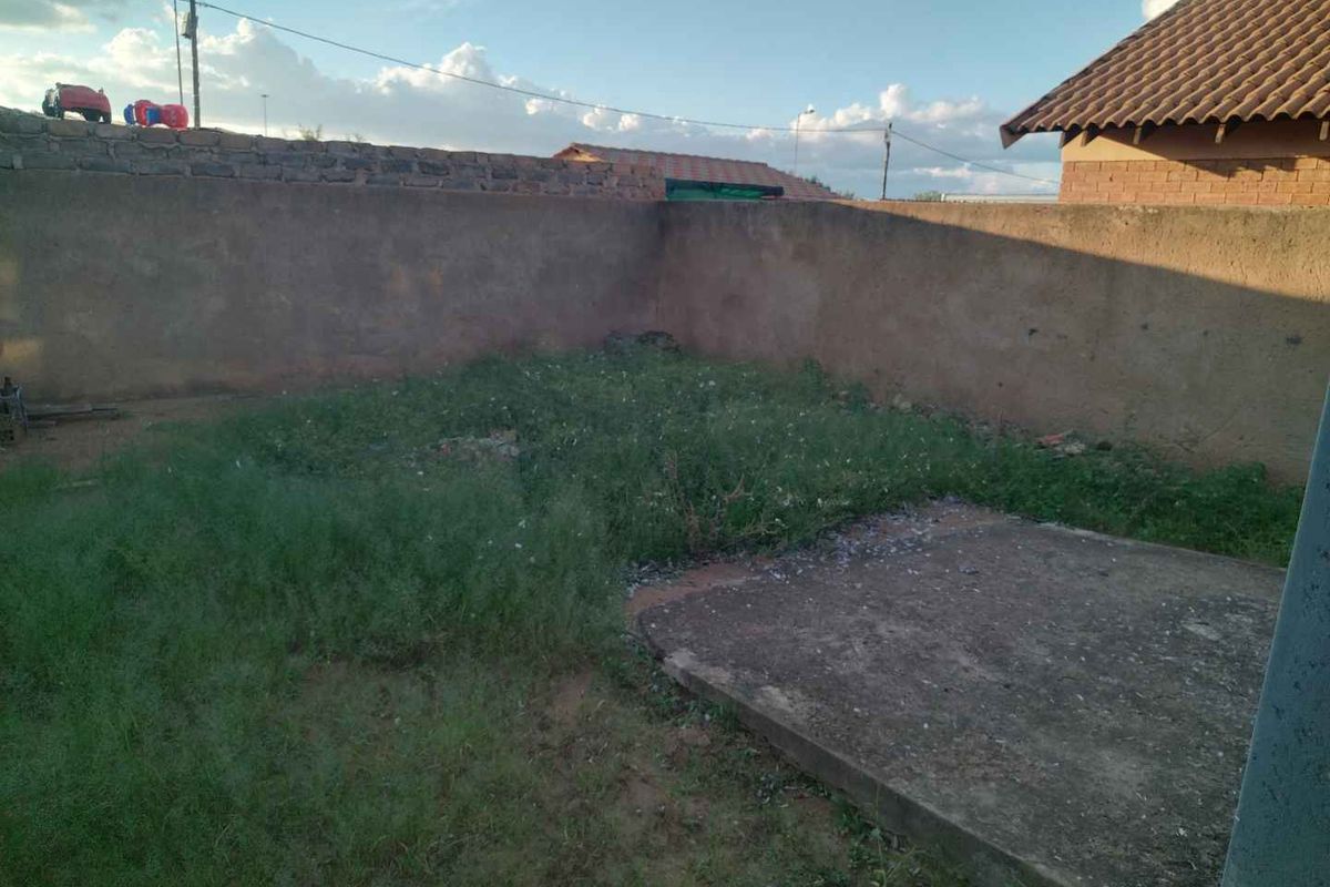 A rear outdoor area featuring a concrete slab, perimeter boundary wall, and overgrown grass, offering potential for landscaping and garden development.