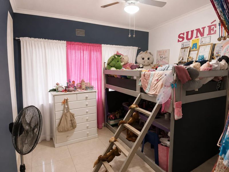 A bedroom featuring tiled flooring, a ceiling fan, and a large window with curtain rails, providing a functional space for a child's room.