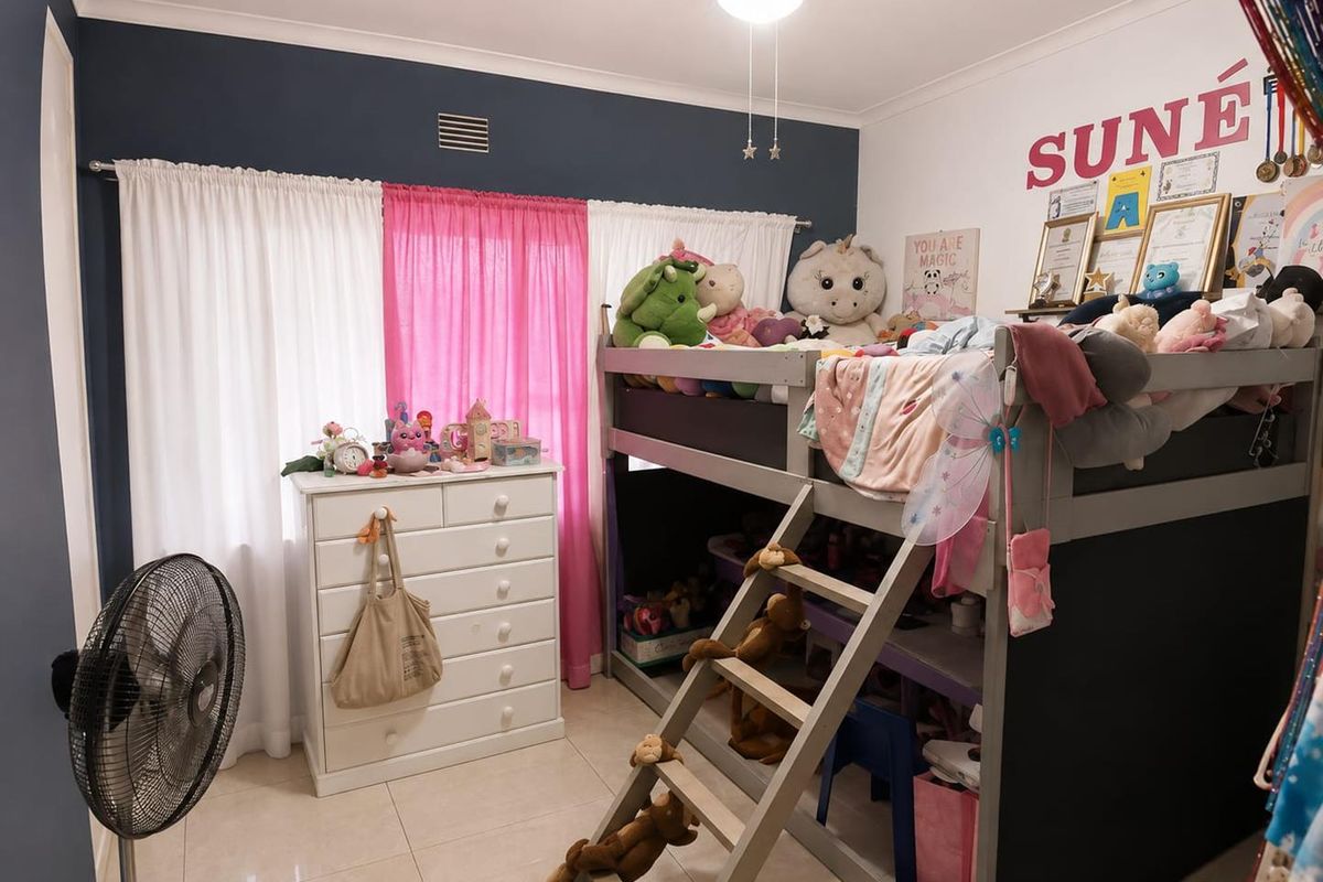 A bedroom featuring tiled flooring, a ceiling fan, and a large window with curtain rails, providing a functional space for a child's room.