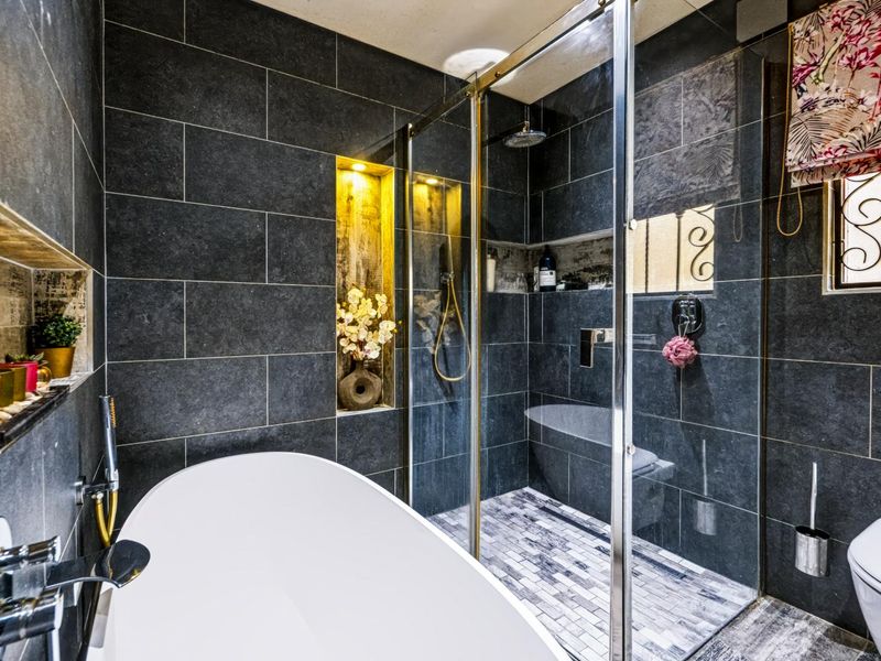 Modern bathroom featuring dark tiled walls, a sleek freestanding bathtub, a spacious walk-in shower with glass enclosure, and elegant recessed wall lighting.