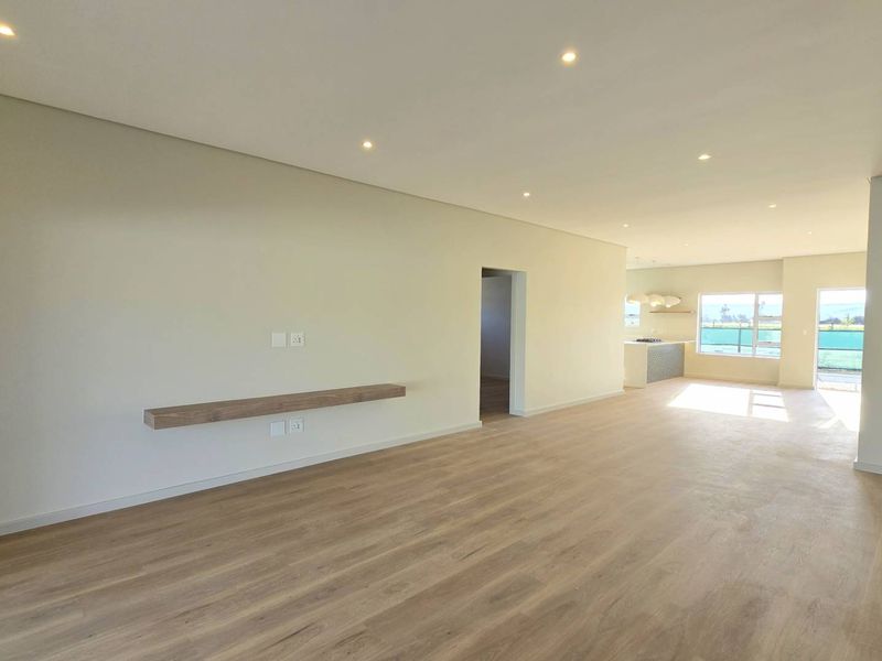 Spacious open-plan living area featuring light laminate flooring, recessed downlighting, and a modern kitchen visible in the background.
