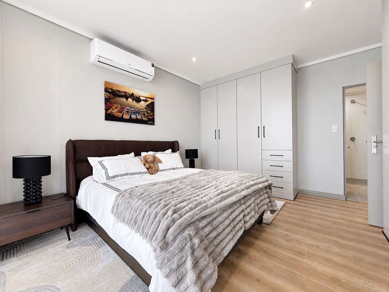 Modern bedroom featuring light laminate flooring, ample built-in cupboard storage, a wall-mounted air conditioning unit, and direct access to an en-suite bathroom.
