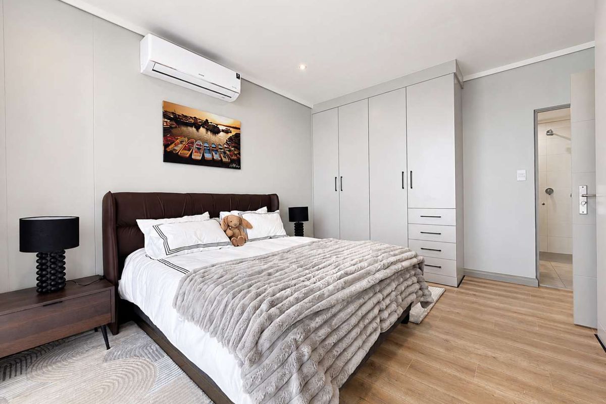 Modern bedroom featuring light laminate flooring, ample built-in cupboard storage, a wall-mounted air conditioning unit, and direct access to an en-suite bathroom.
