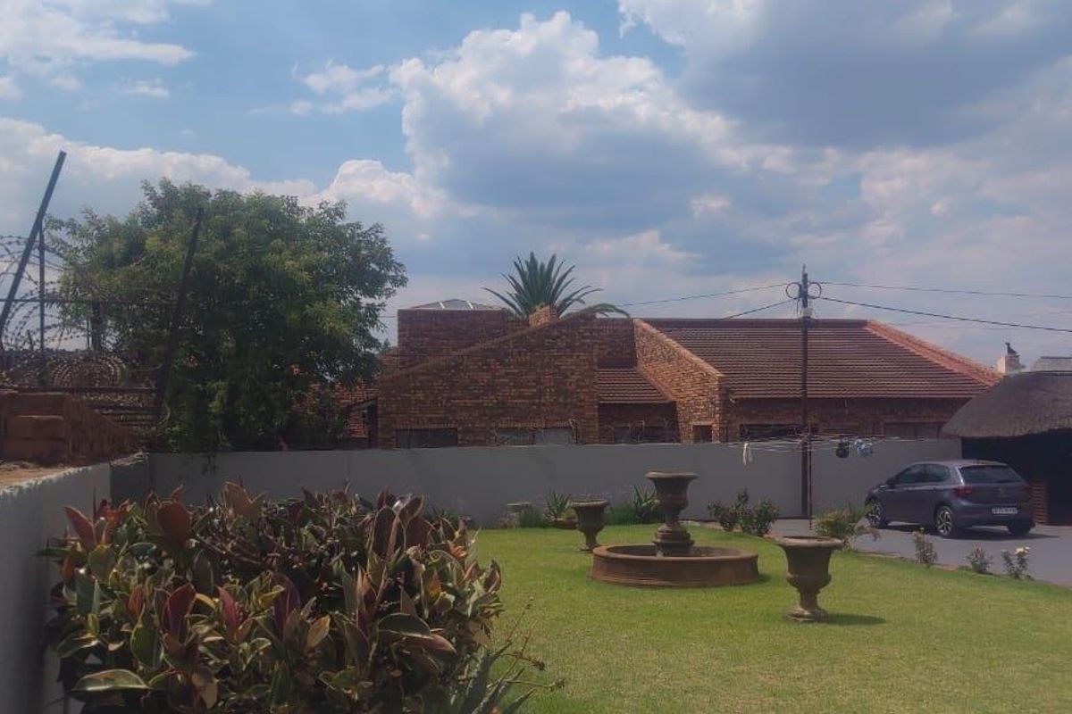A residential garden area featuring a central stone fountain, manicured lawn, and perimeter boundary walling with security fencing.