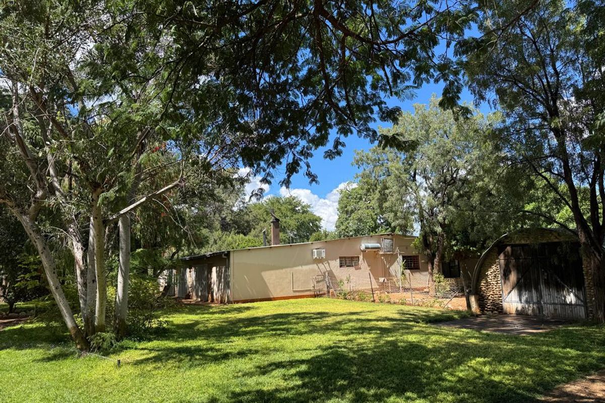 A view of the rear farm property featuring a lawn, mature trees, a single-story building with a chimney, and a detached outbuilding with wooden doors.