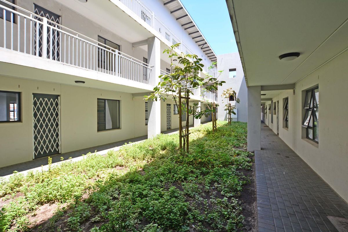 A well-maintained apartment complex courtyard featuring a paved walkway, landscaped garden beds, and access to upper-level balconies with metal security railings.