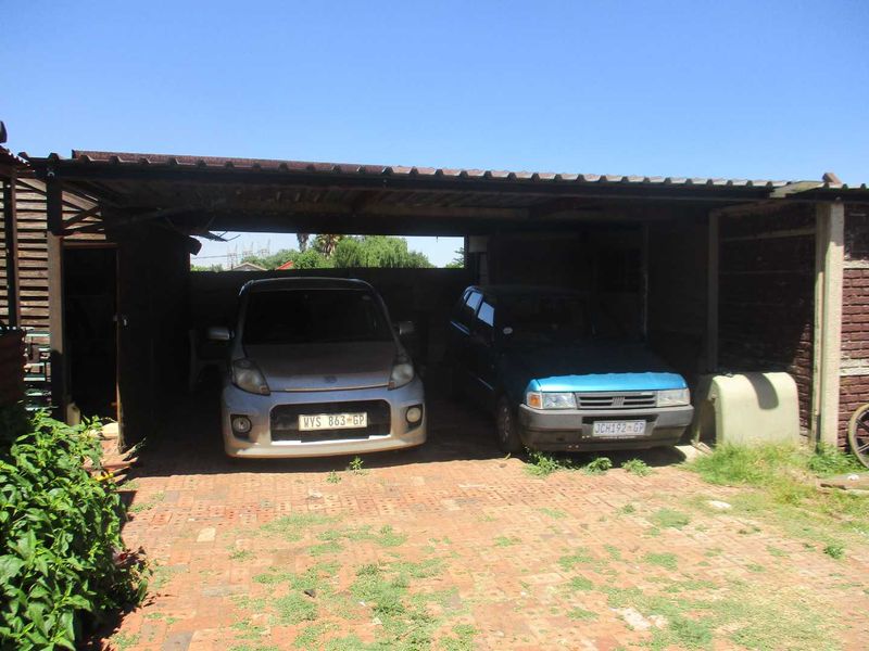 A basic open-sided carport structure with a corrugated metal roof and brick paved flooring, showing signs of wear and maintenance requirements.