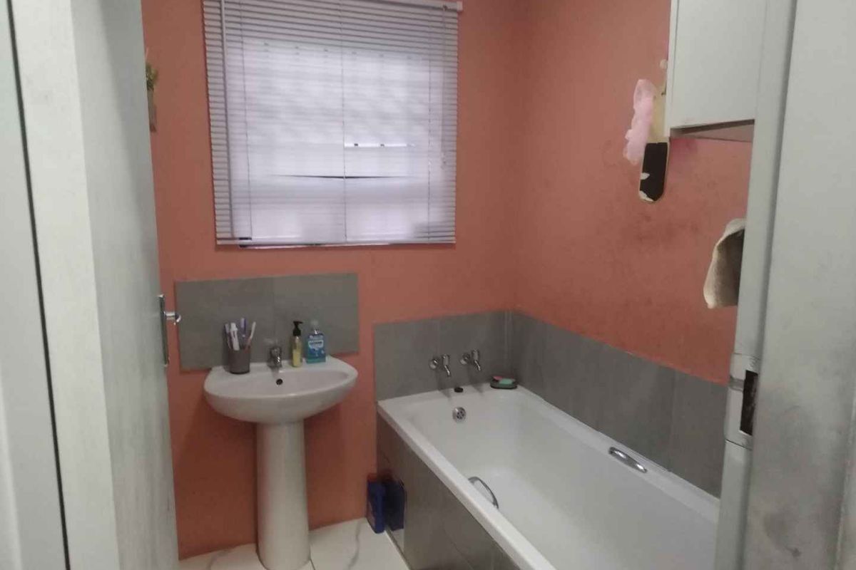 A functional bathroom featuring a pedestal basin, bathtub, and tiled splashback, finished with a neutral venetian blind.