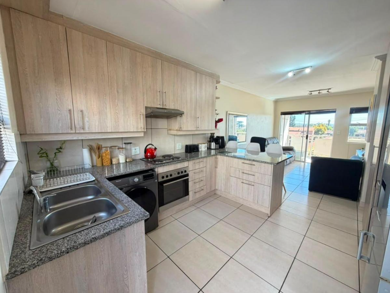 Open-plan kitchen and living area featuring light wood cabinetry, granite countertops, built-in appliances, and tiled flooring with access to a balcony.