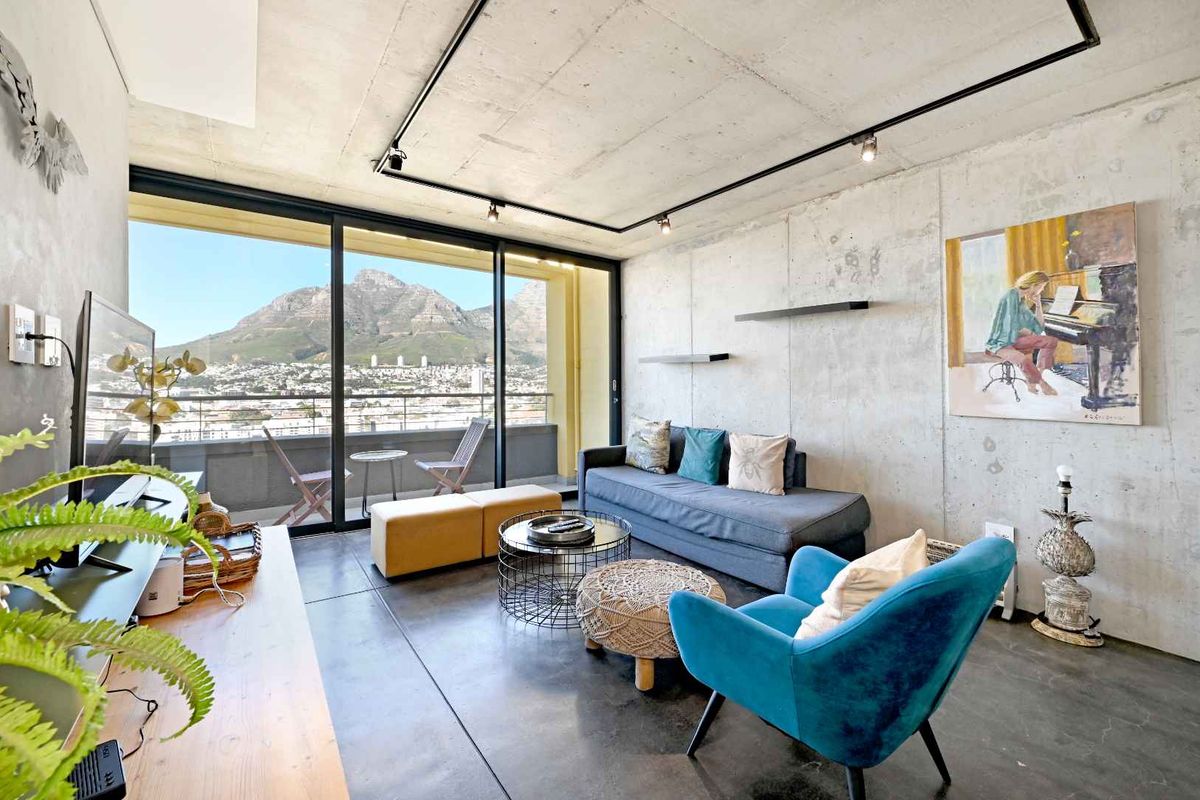 Modern living room featuring industrial-style exposed concrete walls and ceilings, polished concrete floors, and large sliding doors opening to a scenic mountain view balcony.