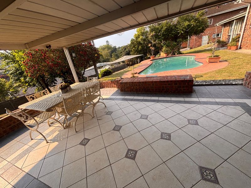 A spacious covered patio featuring decorative tiled flooring, overlooking a private swimming pool and landscaped garden area.