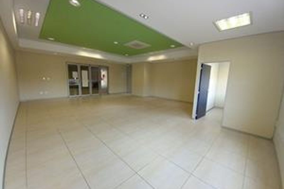 A spacious, empty office area featuring tiled flooring, a dropped ceiling with recessed lighting, and a glass partition door.