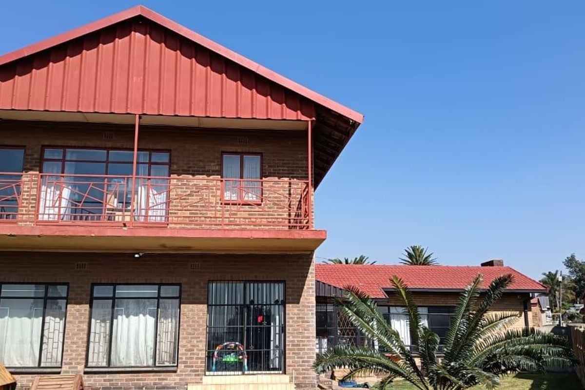 Rear view of a two-story brick house featuring a red metal roof, an upper-level balcony with metal railings, and security bars on windows and doors.