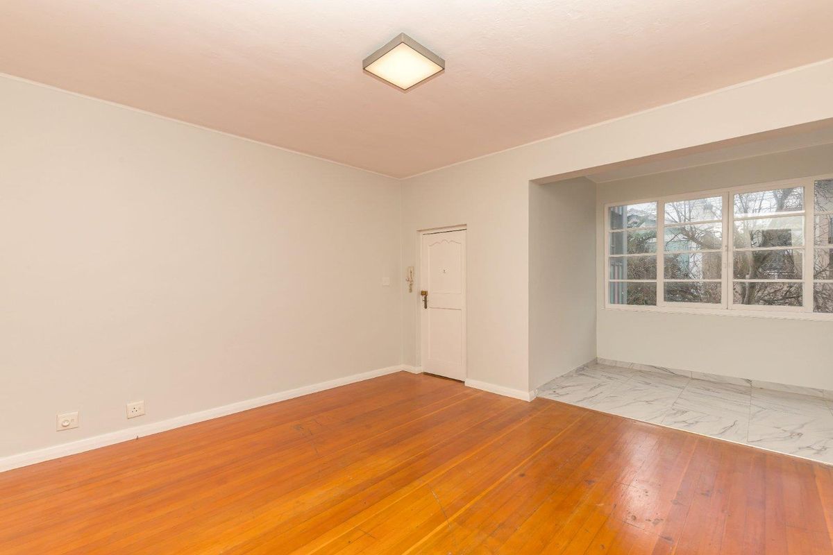 An empty living room featuring polished hardwood flooring, a distinct marble-tiled alcove area, and a bright, neutral interior finish.