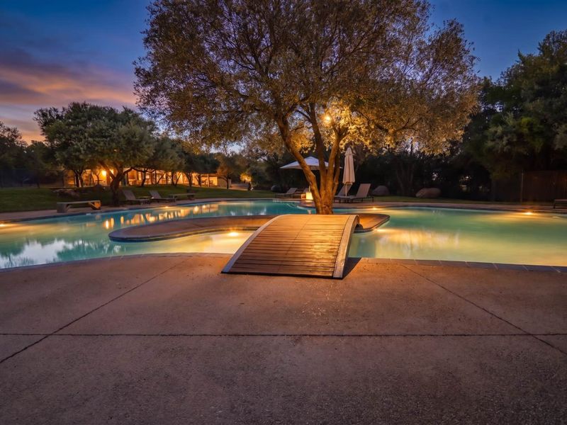 A beautifully illuminated swimming pool area at dusk, featuring a central wooden footbridge and landscaped surroundings.