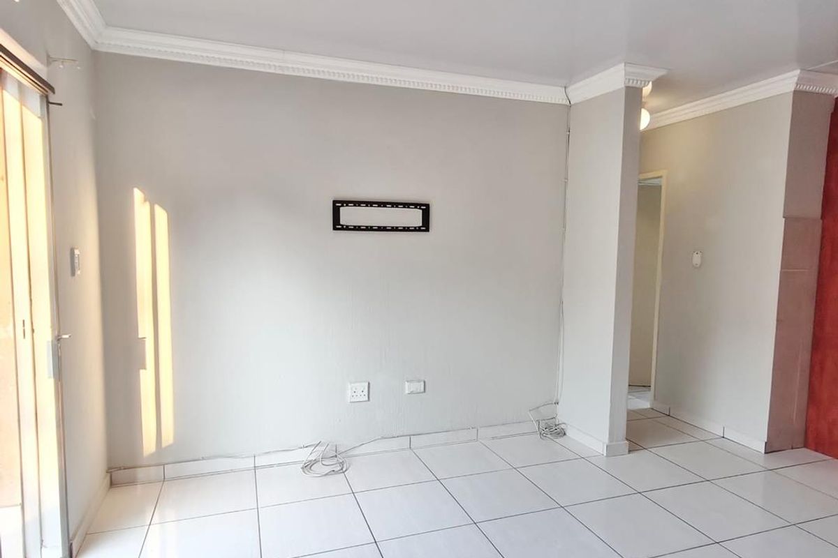A bright, empty living room featuring white tiled flooring, ceiling cornices, and a sliding glass door providing natural light.
