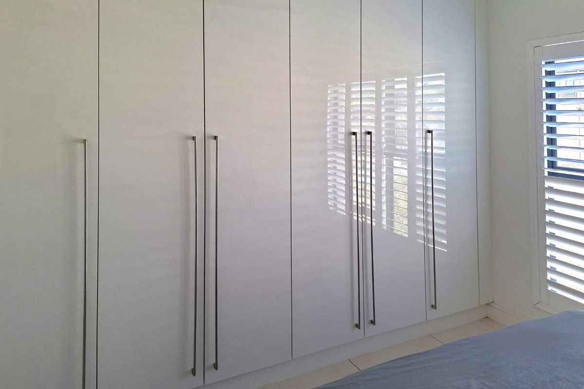 A bedroom featuring modern, floor-to-ceiling white built-in cupboards with sleek handles and white plantation shutters.