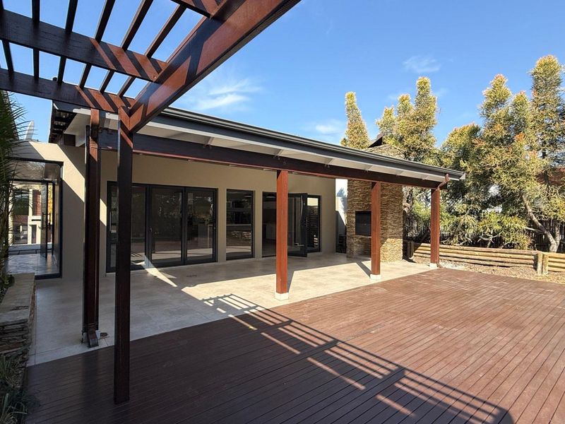 Spacious covered patio featuring timber decking, a pergola, and a built-in stone fireplace, seamlessly connecting to the interior through large stacking glass doors.