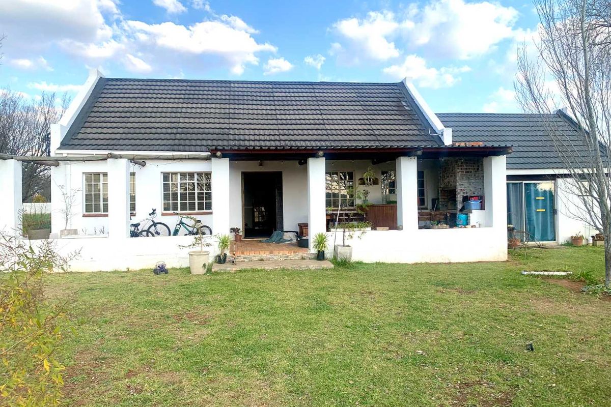 A charming rear exterior view of a small holding residence featuring a covered patio, built-in braai area, and a spacious garden.