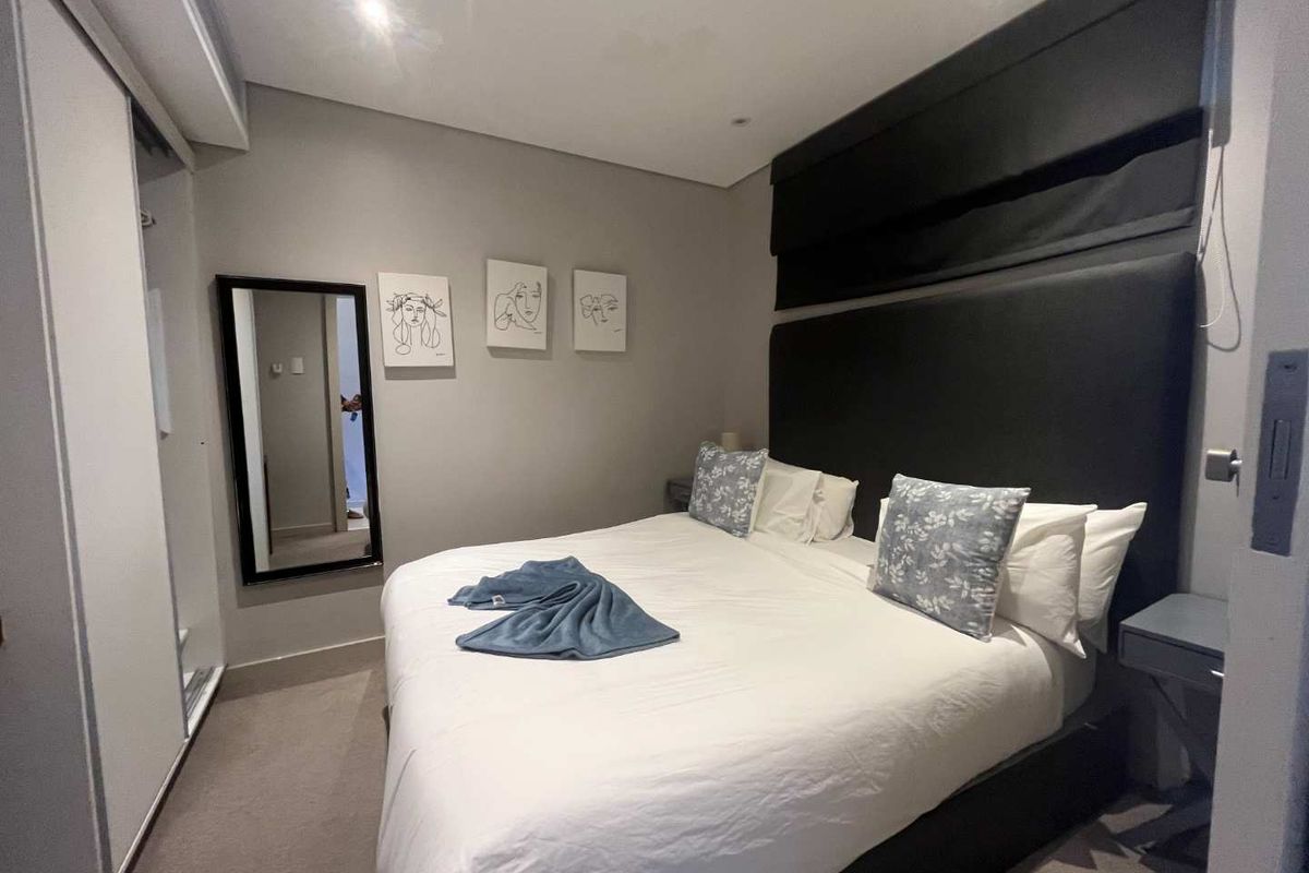 A modern bedroom featuring neutral wall tones, built-in sliding door wardrobes, and a dark upholstered headboard with integrated window blinds.