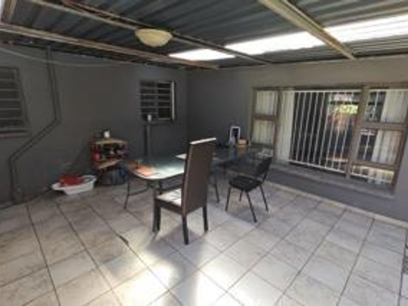 A covered outdoor patio area featuring tiled flooring, security-barred windows, and a corrugated metal roof, offering a functional space for outdoor living.