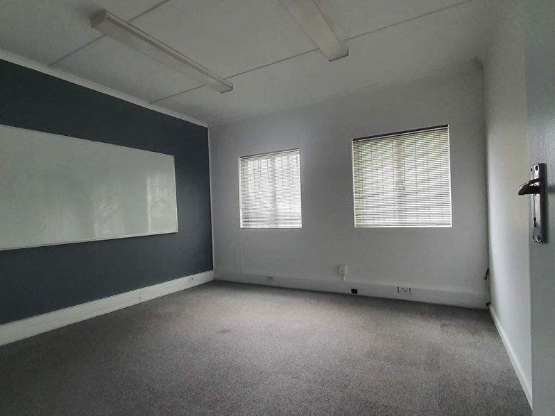 A vacant office space featuring grey carpeting, a wall-mounted whiteboard, dual windows with venetian blinds, and overhead fluorescent lighting.