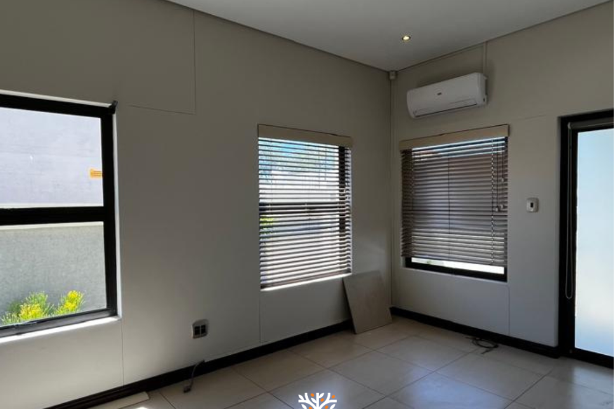 A bright, empty living room featuring tiled flooring, wall-mounted air conditioning unit, and fitted window blinds.