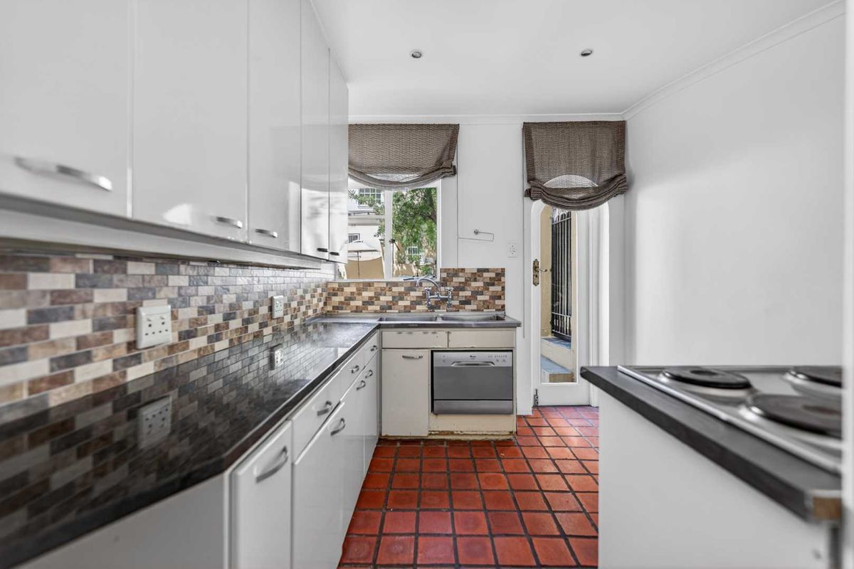A functional kitchen featuring white cabinetry, dark countertops, a mosaic tile backsplash, and terracotta floor tiles with direct access to the exterior.