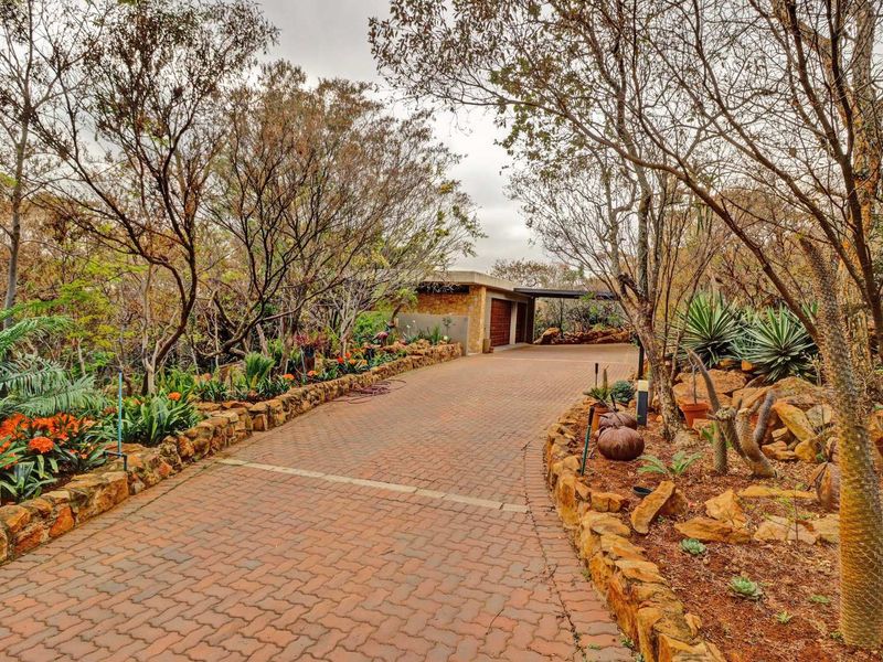 A scenic brick-paved driveway leads to a double garage, framed by mature trees and natural stone-walled garden beds.